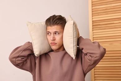 Annoyed young man covering his ears with pillows due to loud sound at home