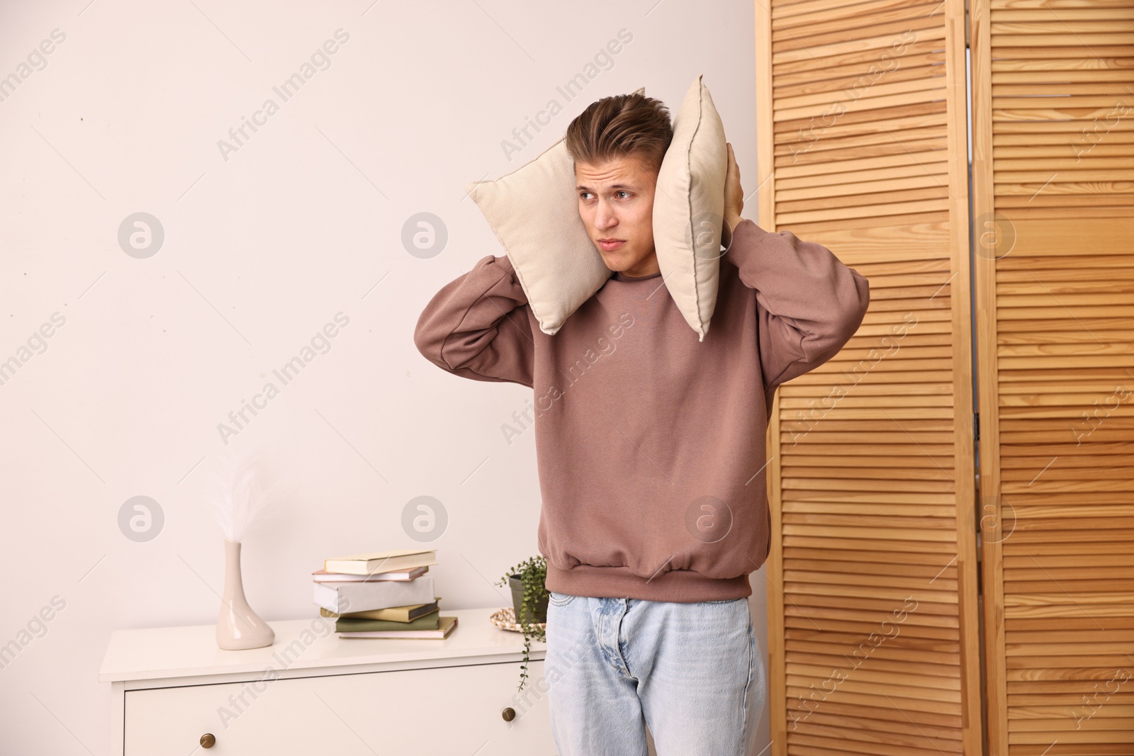 Photo of Annoyed young man covering his ears with pillows due to loud sound at home