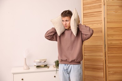 Annoyed young man covering his ears with pillows due to loud sound at home