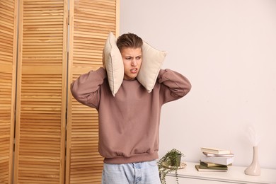 Annoyed young man covering his ears with pillows due to loud sound at home