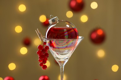 Photo of Creative presentation of Christmas cocktail in bauble and glass against dark beige background with blurred lights, closeup