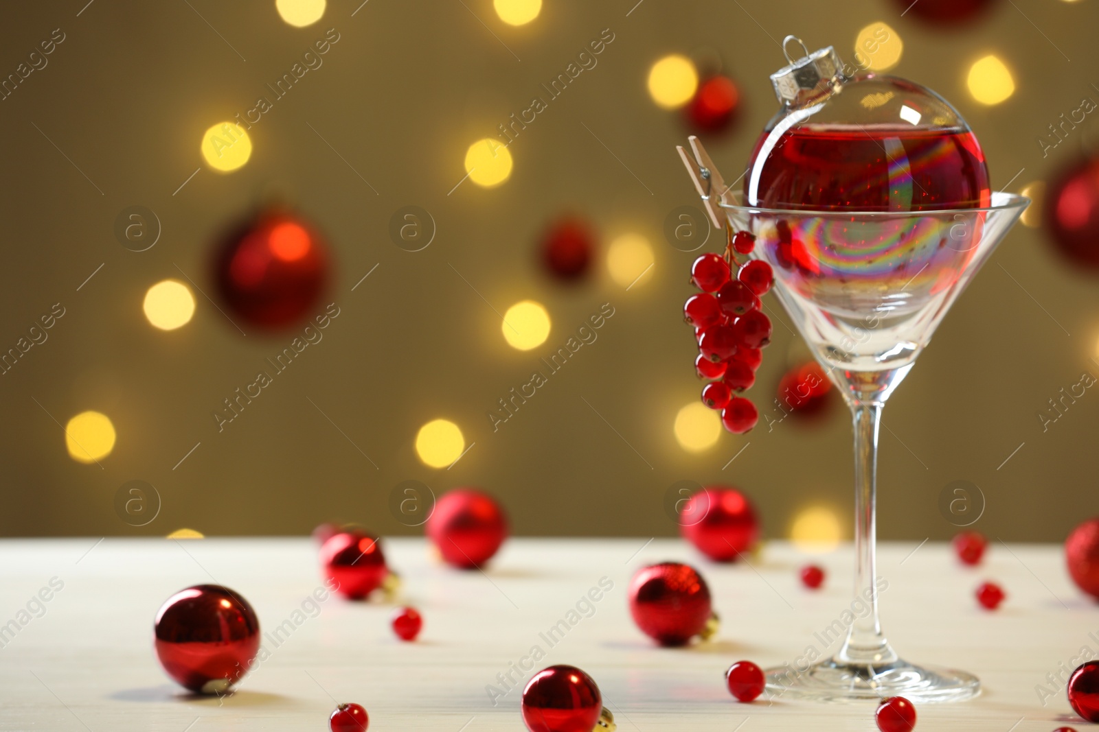 Photo of Creative presentation of Christmas cocktail in bauble and glass on white table against dark beige background with blurred lights. Space for text