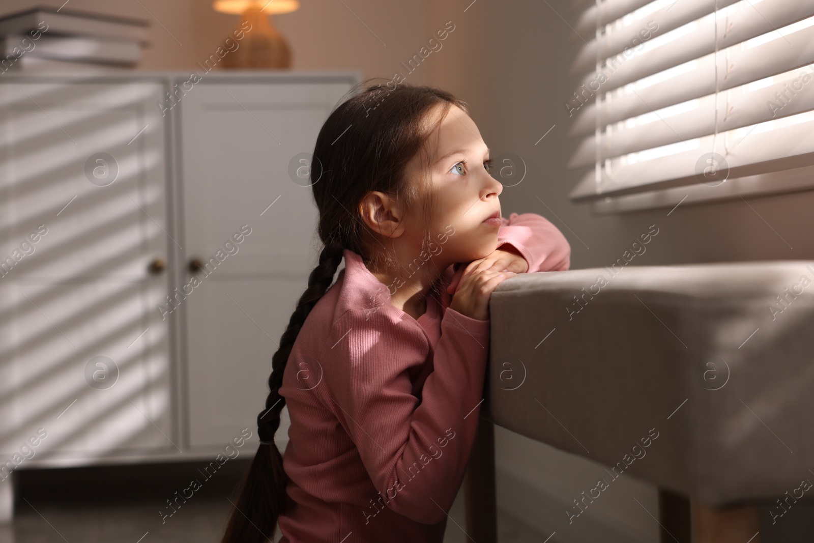 Photo of Orphanage concept. Sad girl and looking through window blinds indoors