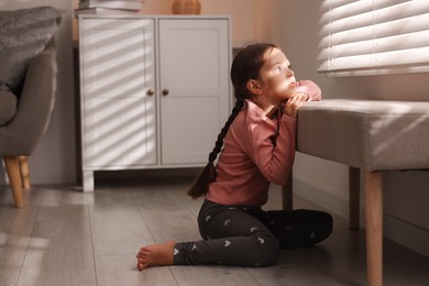 Orphanage concept. Sad girl and looking through window blinds indoors