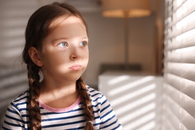 Photo of Orphanage concept. Sad girl and looking through window blinds indoors