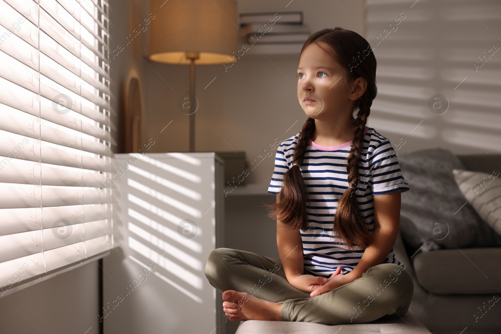 Photo of Orphanage concept. Sad girl sitting on bench and looking through window blinds indoors