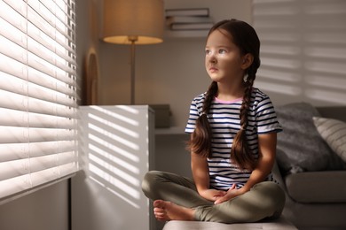 Orphanage concept. Sad girl sitting on bench and looking through window blinds indoors