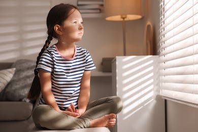 Photo of Orphanage concept. Sad girl sitting on bench and looking through window blinds indoors