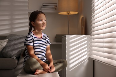 Photo of Orphanage concept. Sad girl sitting on bench and looking through window blinds indoors