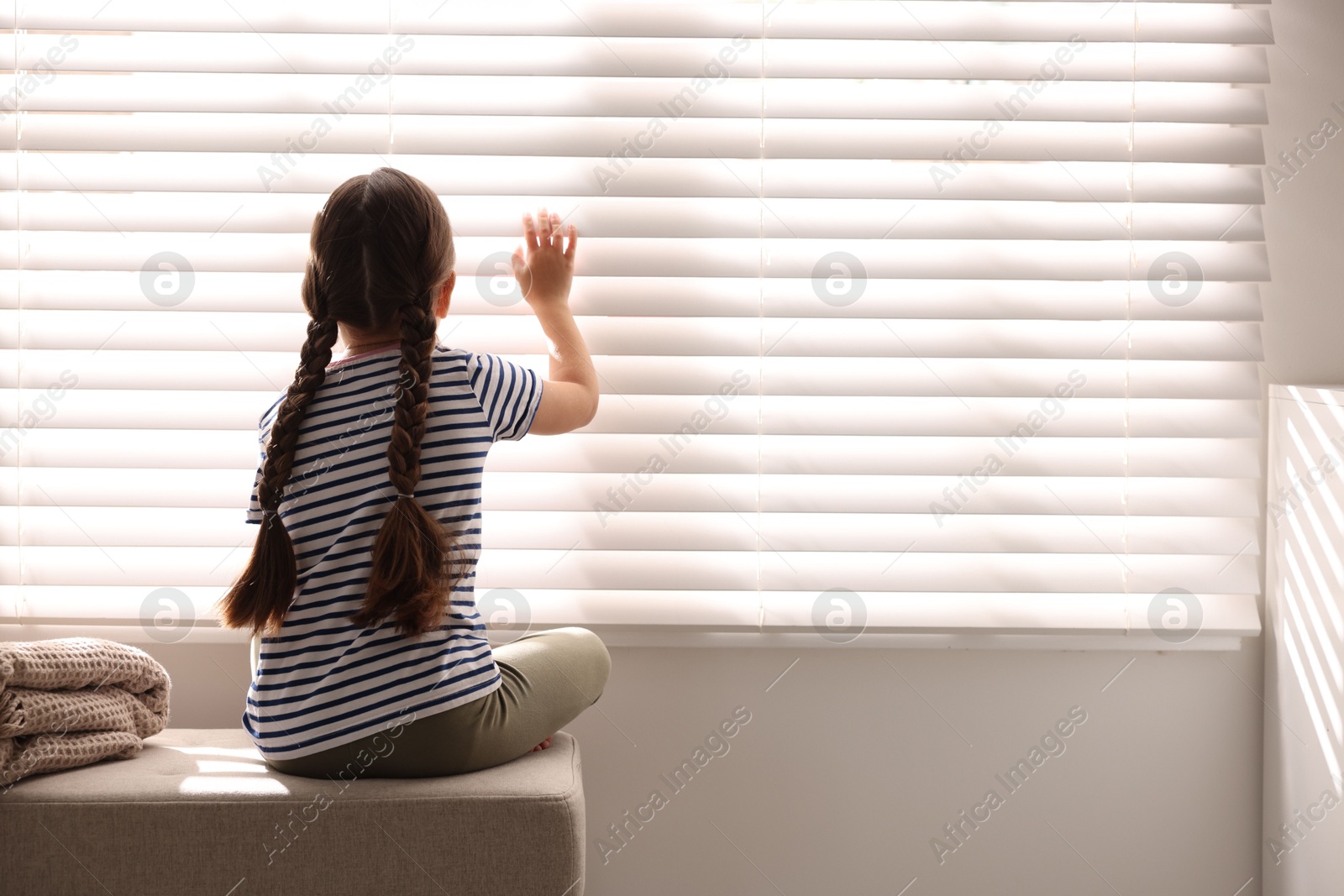 Photo of Orphanage concept. Girl sitting on bench and looking through window blinds indoors, back view