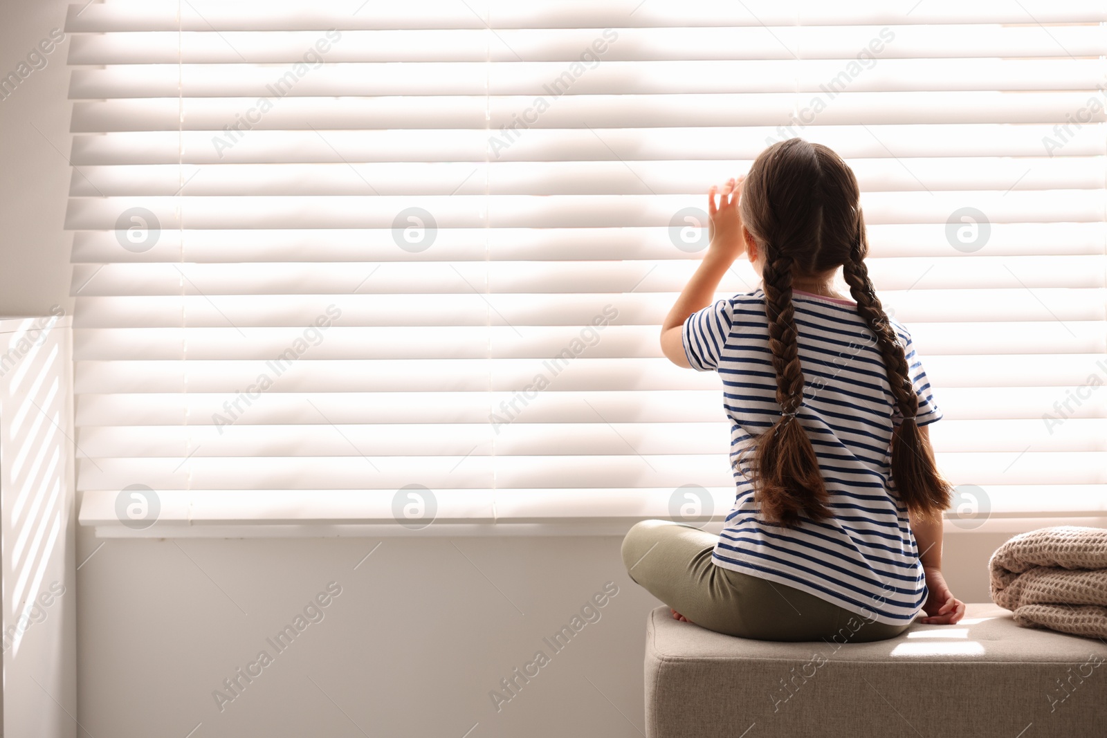 Photo of Orphanage concept. Girl sitting on bench and looking through window blinds indoors, back view