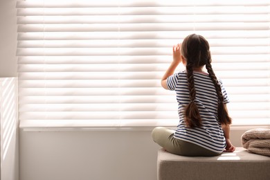 Orphanage concept. Girl sitting on bench and looking through window blinds indoors, back view