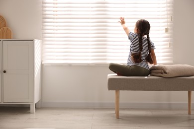 Photo of Orphanage concept. Girl sitting on bench and looking through window blinds indoors, back view