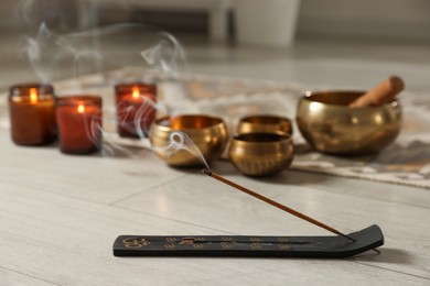 Photo of Incense stick smoldering in holder, burning candles and tibetan singing bowls on floor, selective focus