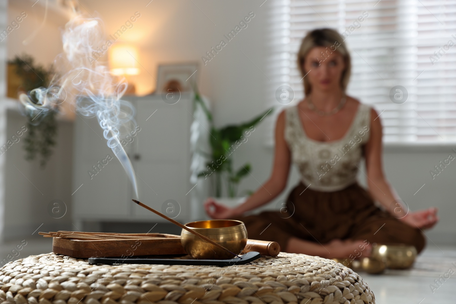 Photo of Young woman practicing yoga indoors, focus on smoldering incense stick and tibetan singing bowl