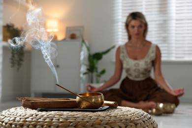 Photo of Young woman practicing yoga indoors, focus on smoldering incense stick and tibetan singing bowl