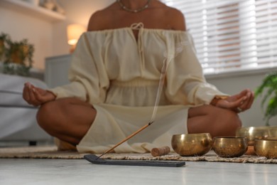 Photo of Young woman with smoldering incense stick and tibetan singing bowls practicing yoga on floor indoors, selective focus