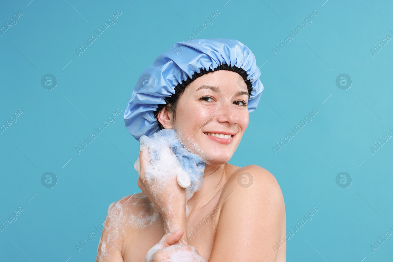 Photo of Woman in shower cap covered with soap foam holding mesh sponge on light blue background