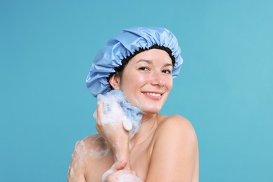 Photo of Woman in shower cap covered with soap foam holding mesh sponge on light blue background