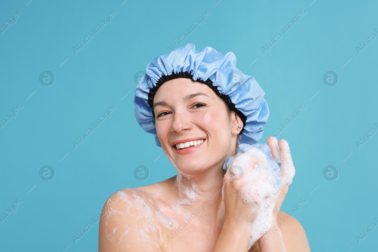 Photo of Woman in shower cap covered with soap foam holding mesh sponge on light blue background