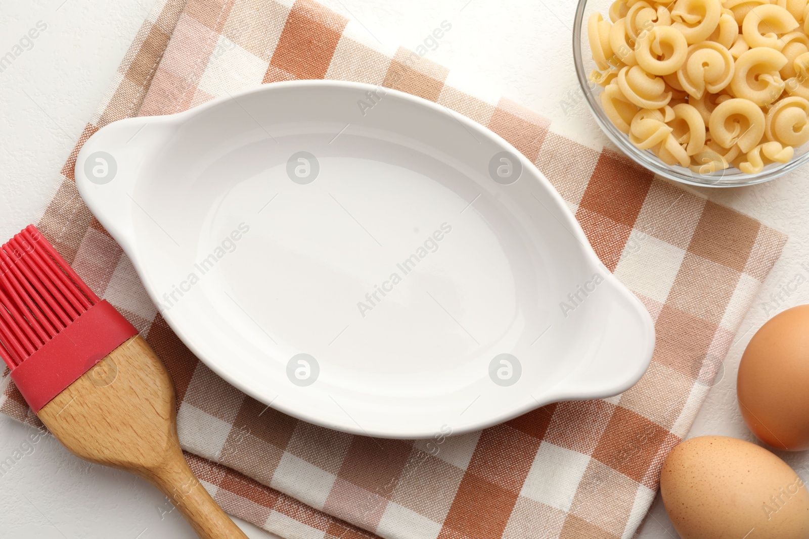 Photo of Ceramic casserole, raw pasta, eggs and brush on white table, flat lay