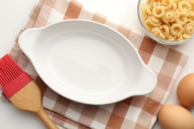 Photo of Ceramic casserole, raw pasta, eggs and brush on white table, flat lay