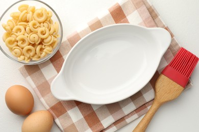 Photo of Ceramic casserole, raw pasta, eggs and brush on white table, flat lay