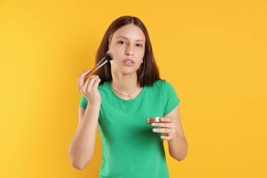 Photo of Teenage girl applying blusher with makeup brush on yellow background