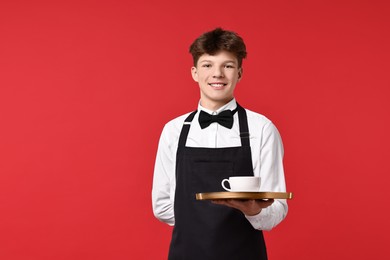 Photo of Teenage boy with cup of drink working as waiter on red background