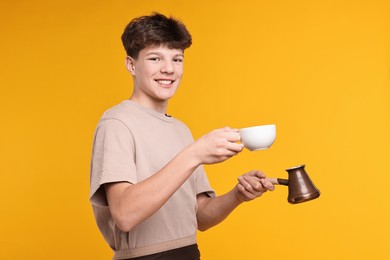 Photo of Teenage boy with cezve pot and cup working as barista on orange background