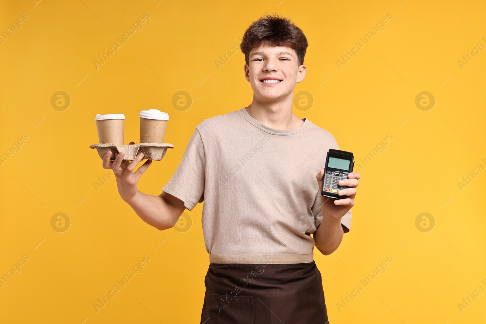 Photo of Teenage boy with paper cups and payment terminal working as barista on orange background