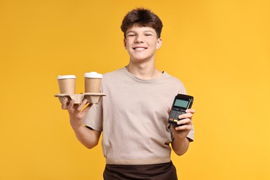 Photo of Teenage boy with paper cups and payment terminal working as barista on orange background