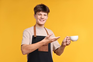 Photo of Teenage boy with cup of coffee working as barista on orange background