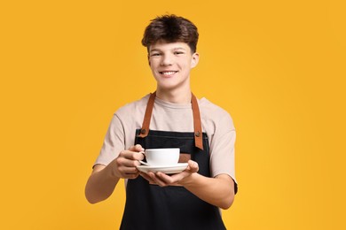 Photo of Teenage boy with cup of coffee working as barista on orange background