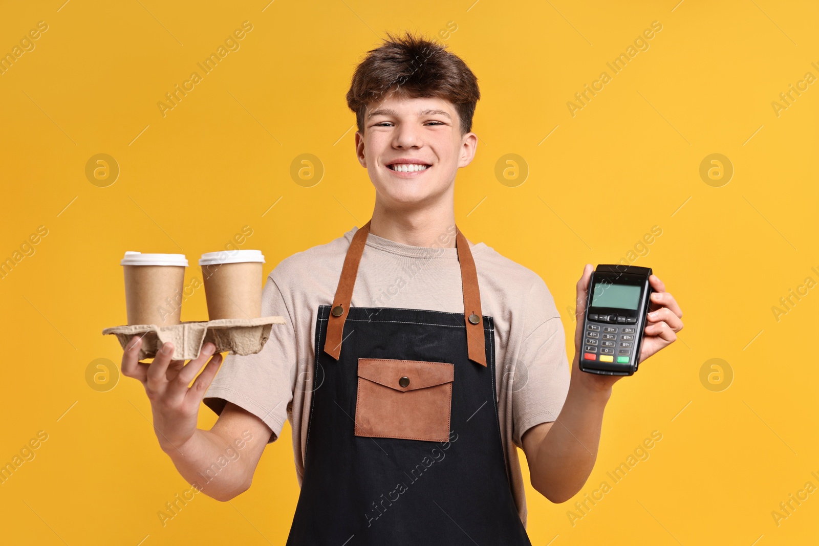 Photo of Teenage boy with paper cups and payment terminal working as barista on orange background