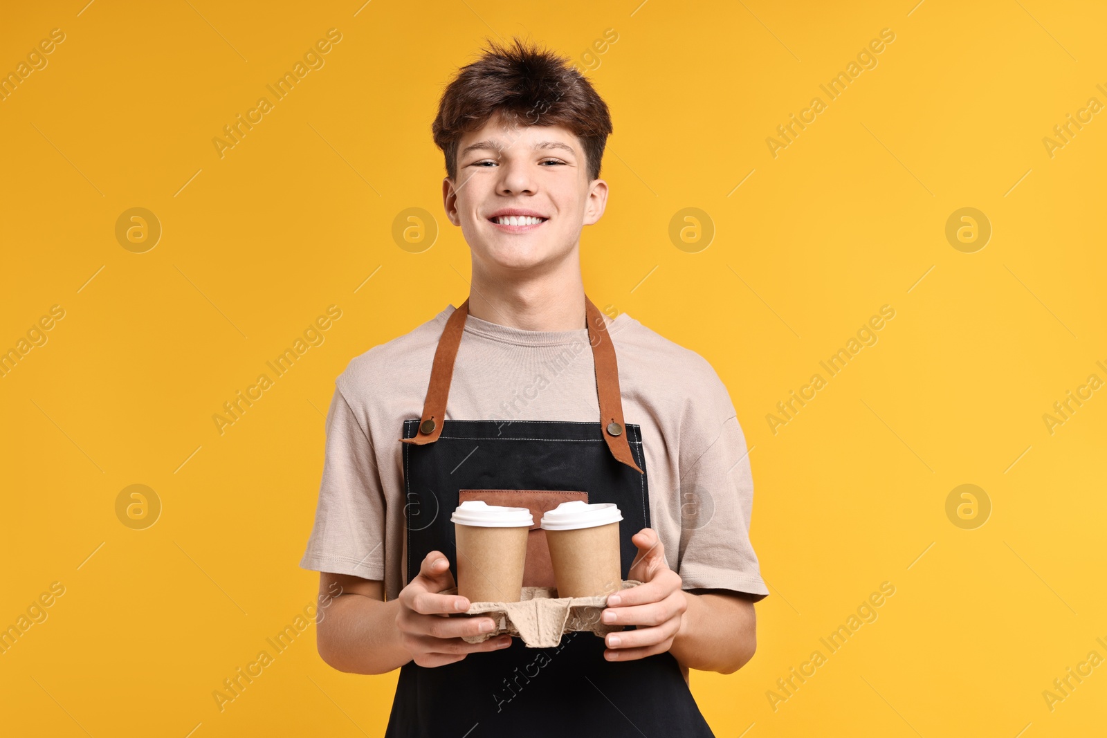 Photo of Teenage boy with paper cups working as barista on orange background