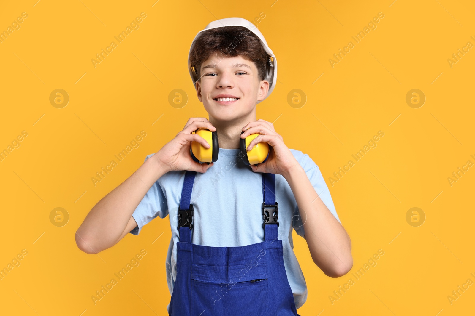 Photo of Teenage boy in hardhat with earmuffs working as builder on orange background