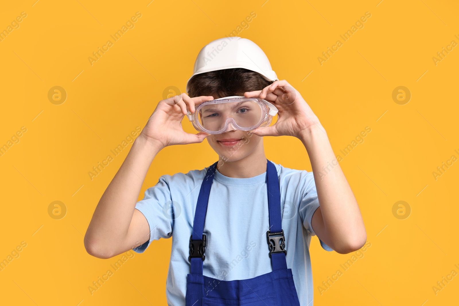 Photo of Teenage boy in hardhat and protective mask working as builder on orange background