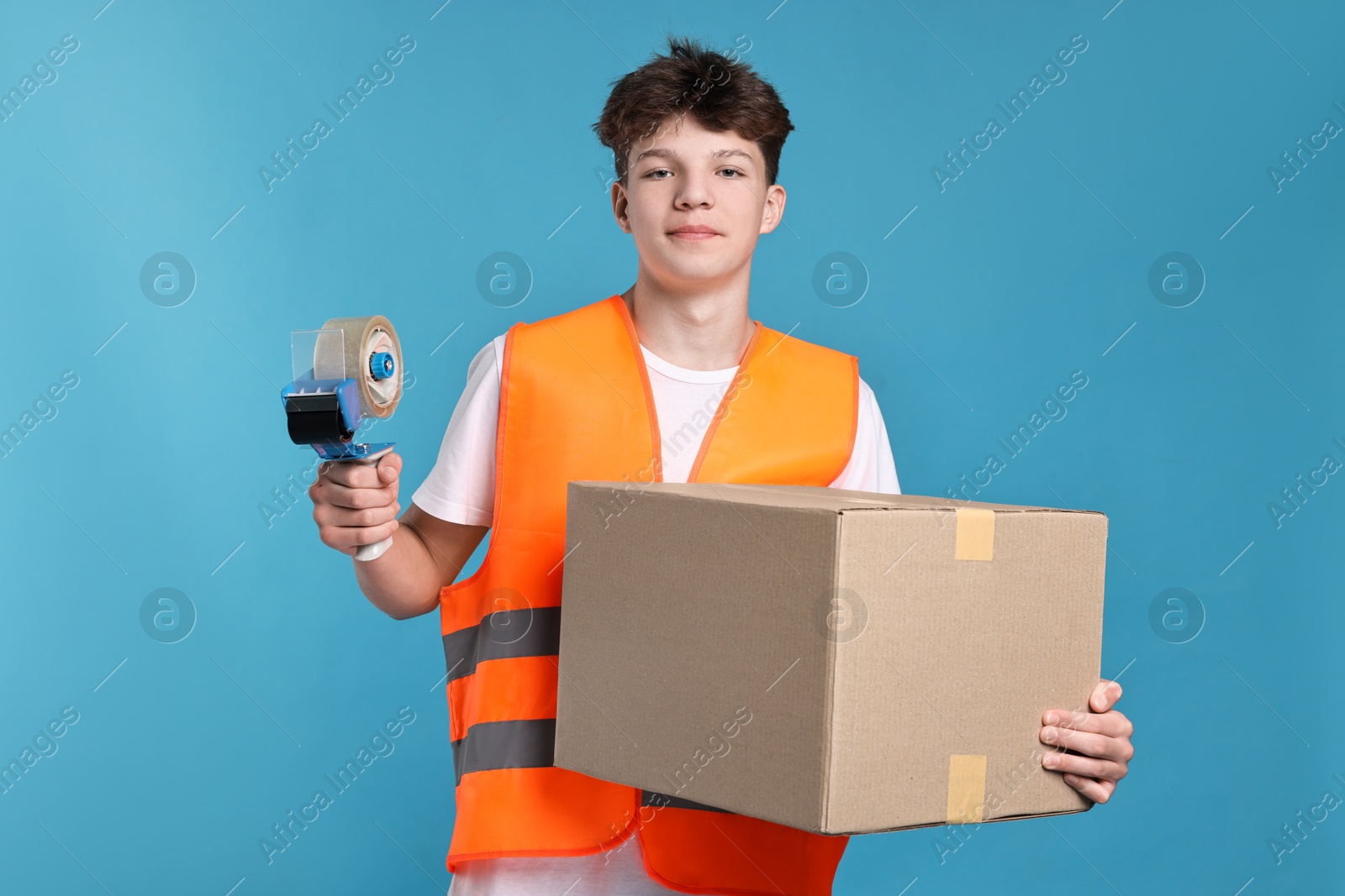 Photo of Teenage boy with tape gun dispenser and box in safety vest working as warehouse packer on blue background