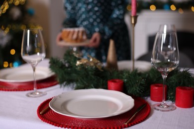 Photo of Woman setting table for Christmas dinner at home, focus on plate, glasses and decor