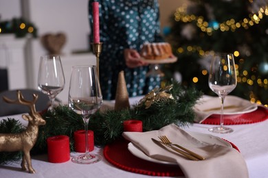 Photo of Woman setting table for Christmas dinner at home, focus on dishware, glasses and decor