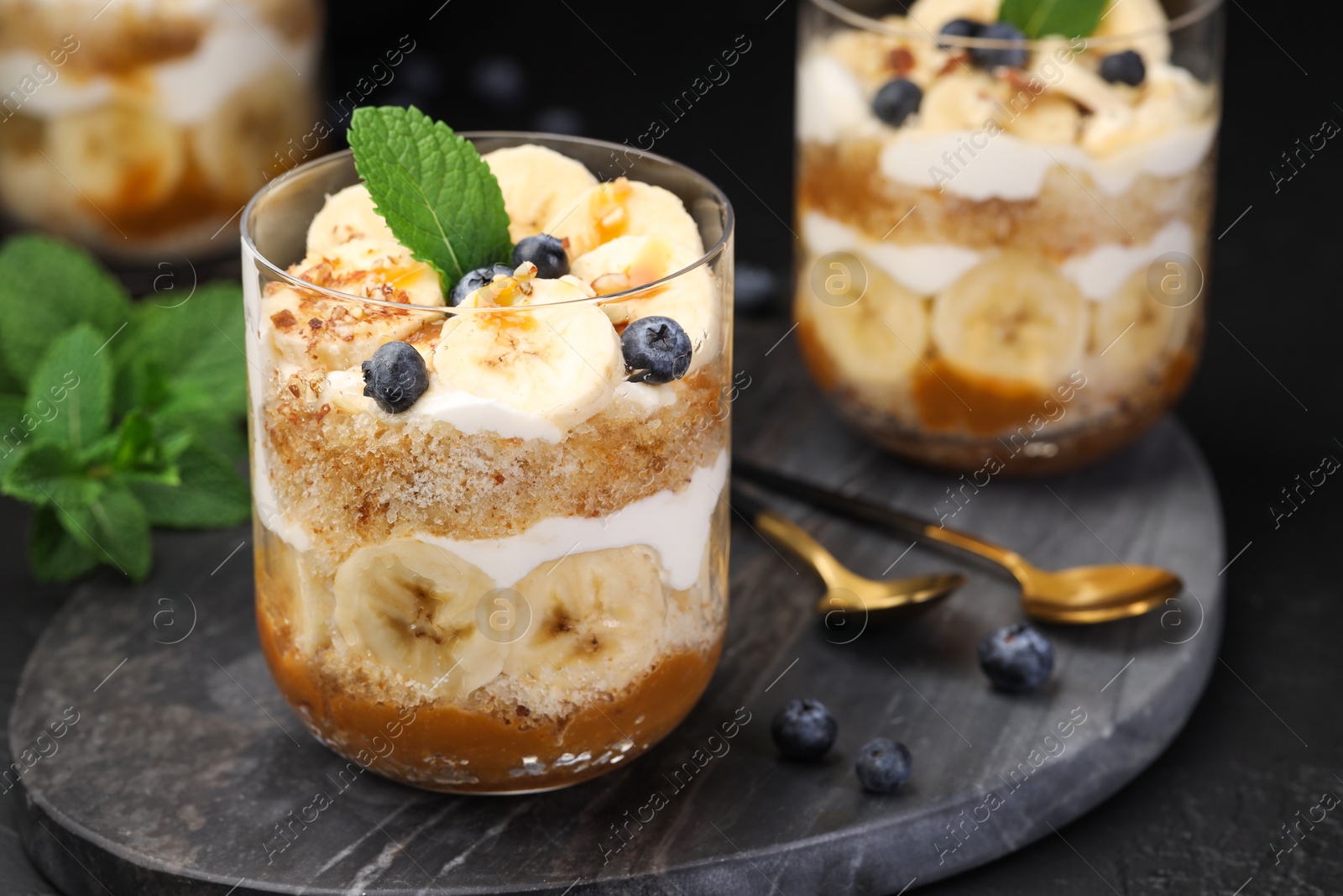 Photo of Tasty trifle dessert. Sponge cake, blueberries, banana and whipped cream in glasses on black table, closeup