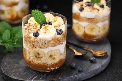 Photo of Tasty trifle dessert. Sponge cake, blueberries, banana and whipped cream in glasses on black table, closeup