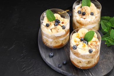 Photo of Tasty trifle dessert. Sponge cake, blueberries, banana and whipped cream in glasses on black table, closeup