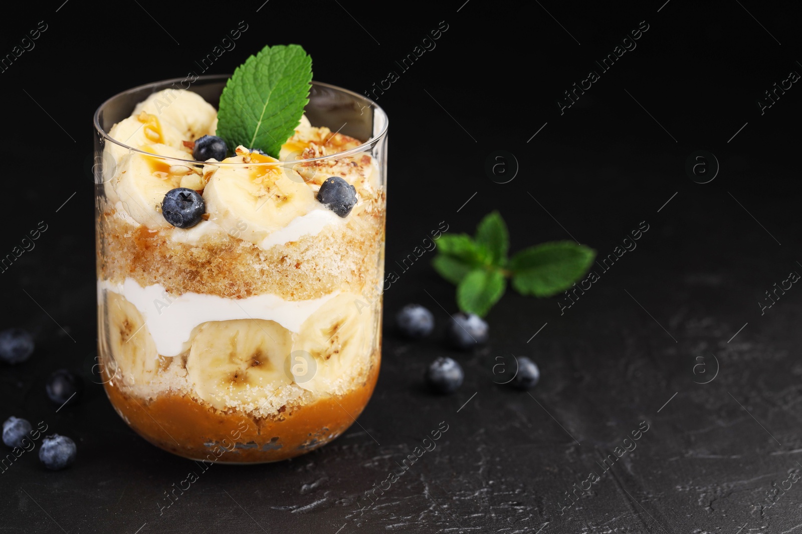 Photo of Tasty trifle dessert. Sponge cake, blueberries, banana and whipped cream in glass on black table, closeup