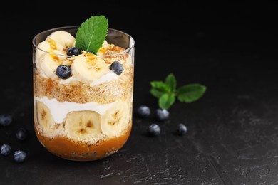 Photo of Tasty trifle dessert. Sponge cake, blueberries, banana and whipped cream in glass on black table, closeup