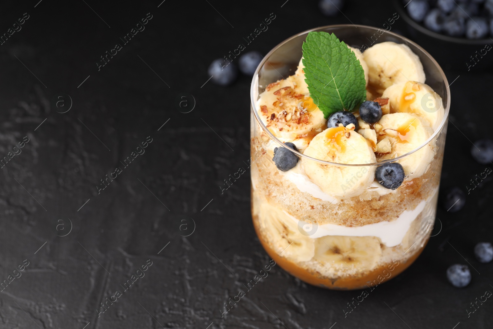 Photo of Tasty trifle dessert. Sponge cake, blueberries, banana and whipped cream in glass on black table, closeup. Space for text