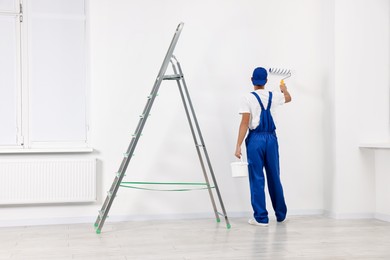 Photo of Male handyman painting wall with roller near ladder indoors, back view