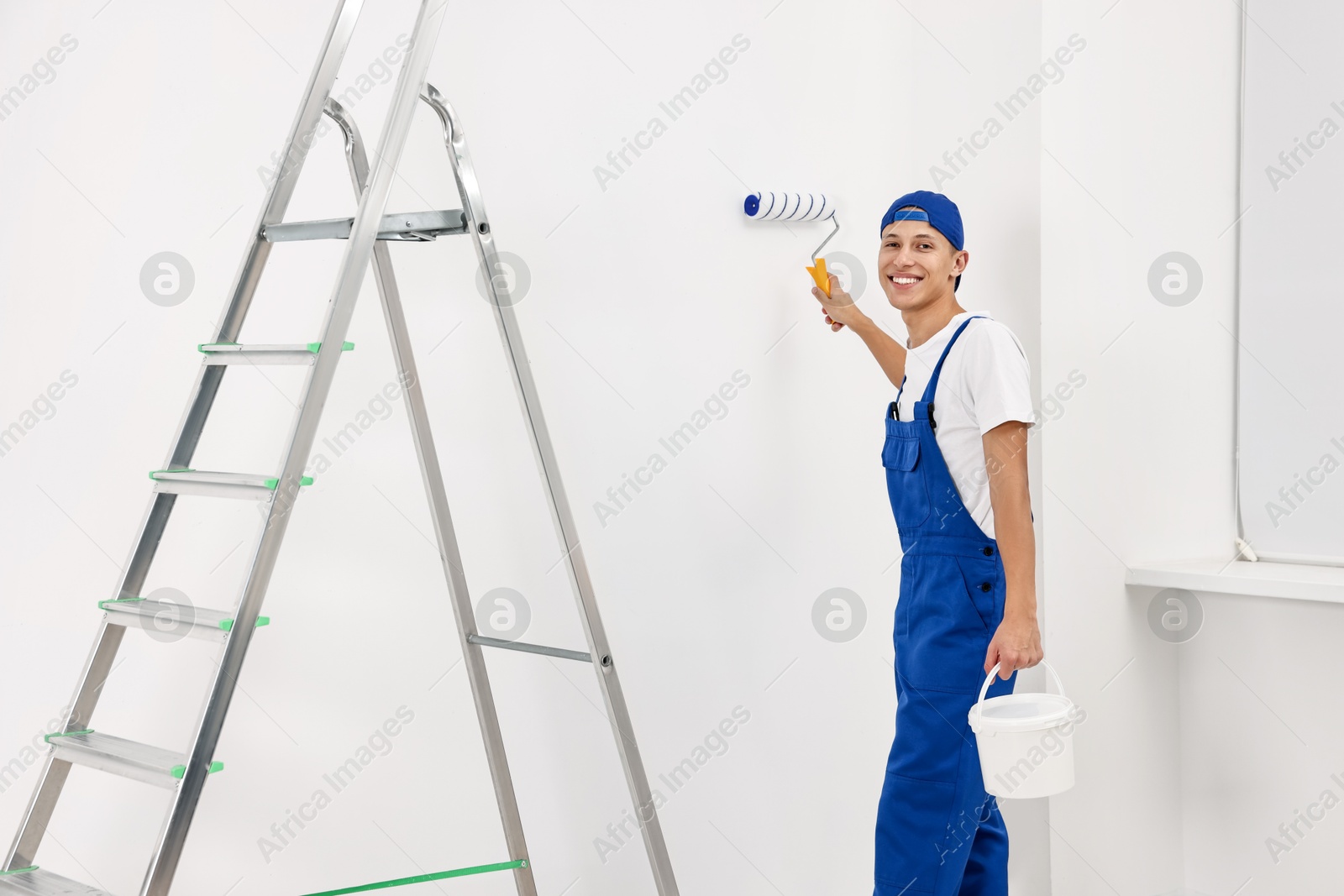 Photo of Smiling handyman painting wall with roller near ladder indoors