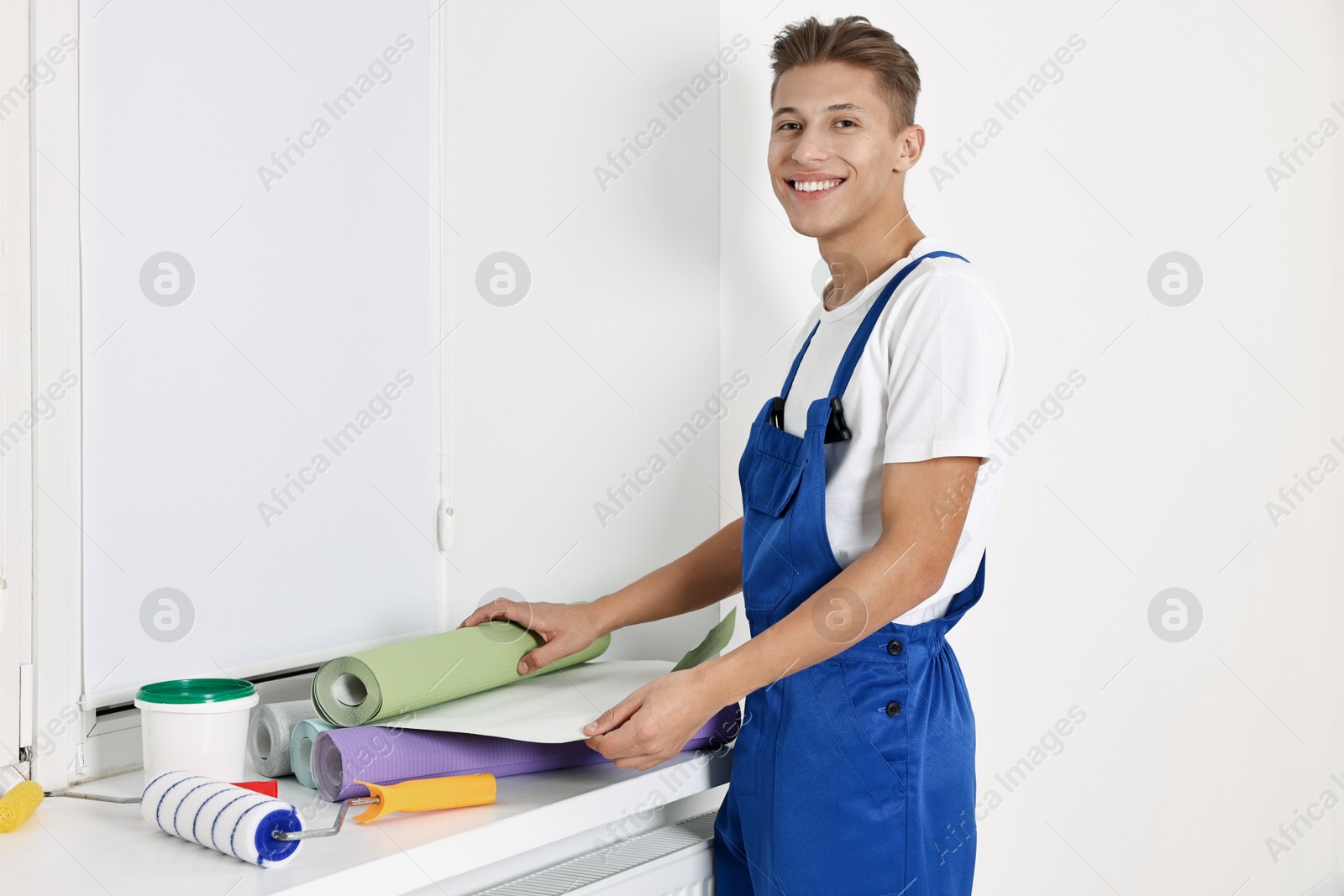 Photo of Smiling handyman with wallpapers and paint roller at window sill indoors